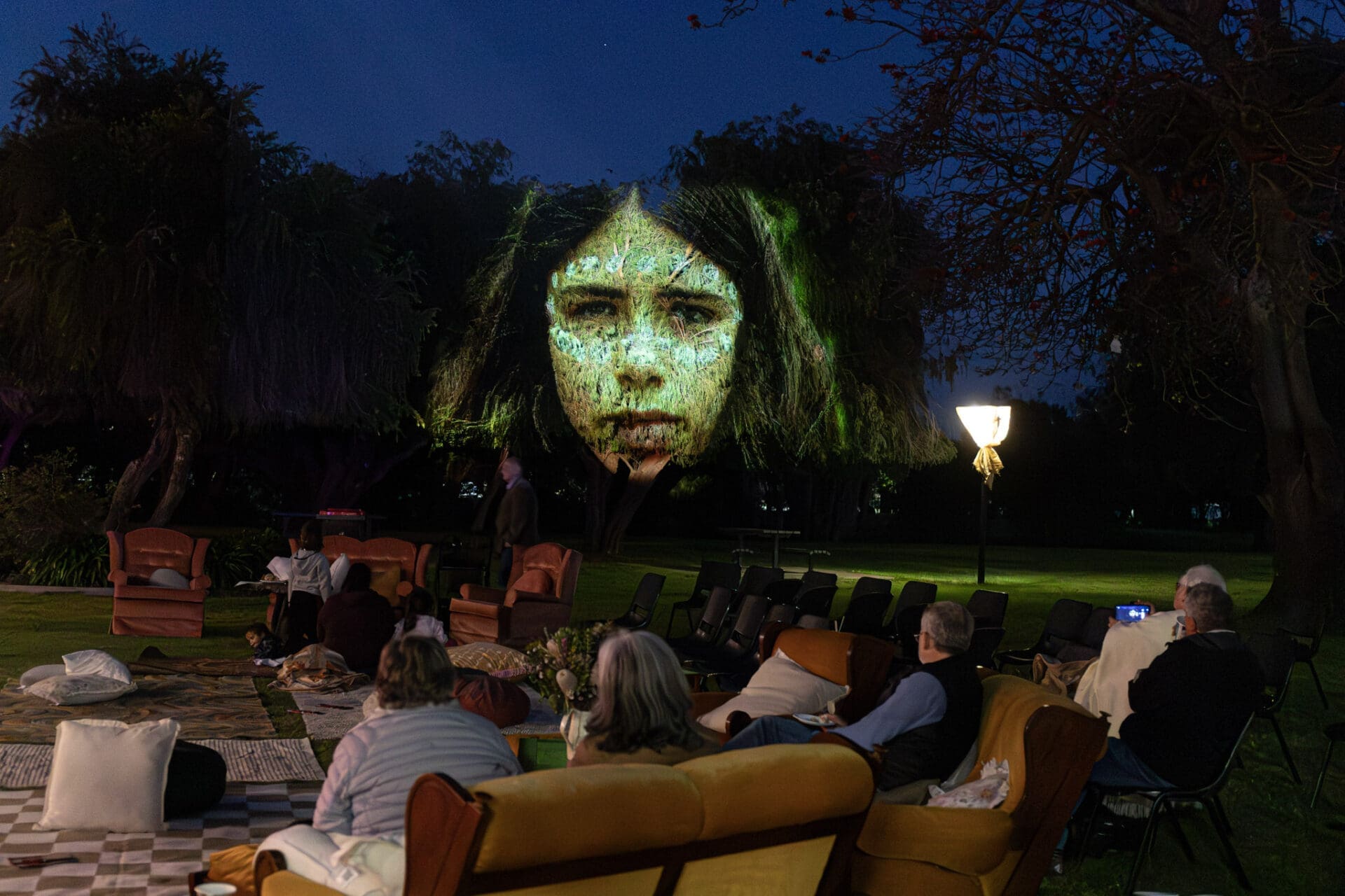 Patrons Sitting Outside At Night Watching A Screen.