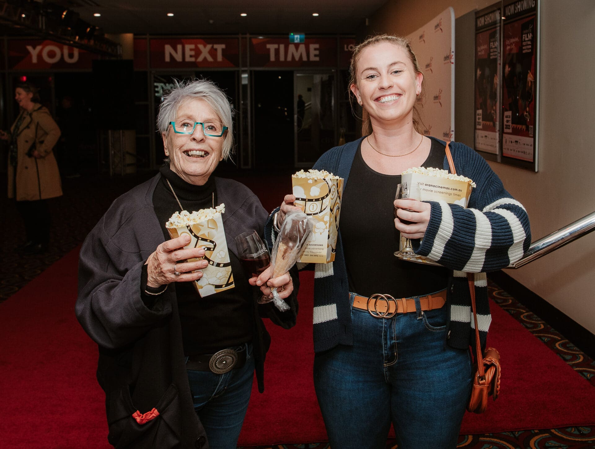 Two Women Smiling At The Camera Holding Popcorn Boxes And Glasses Of Wine Outside The Cinema.